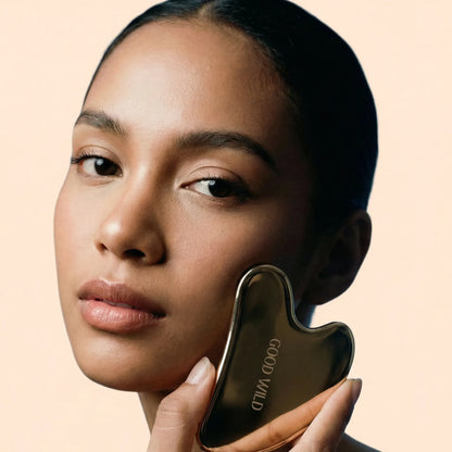 Woman holding a gold facial tool against her face on a beige background