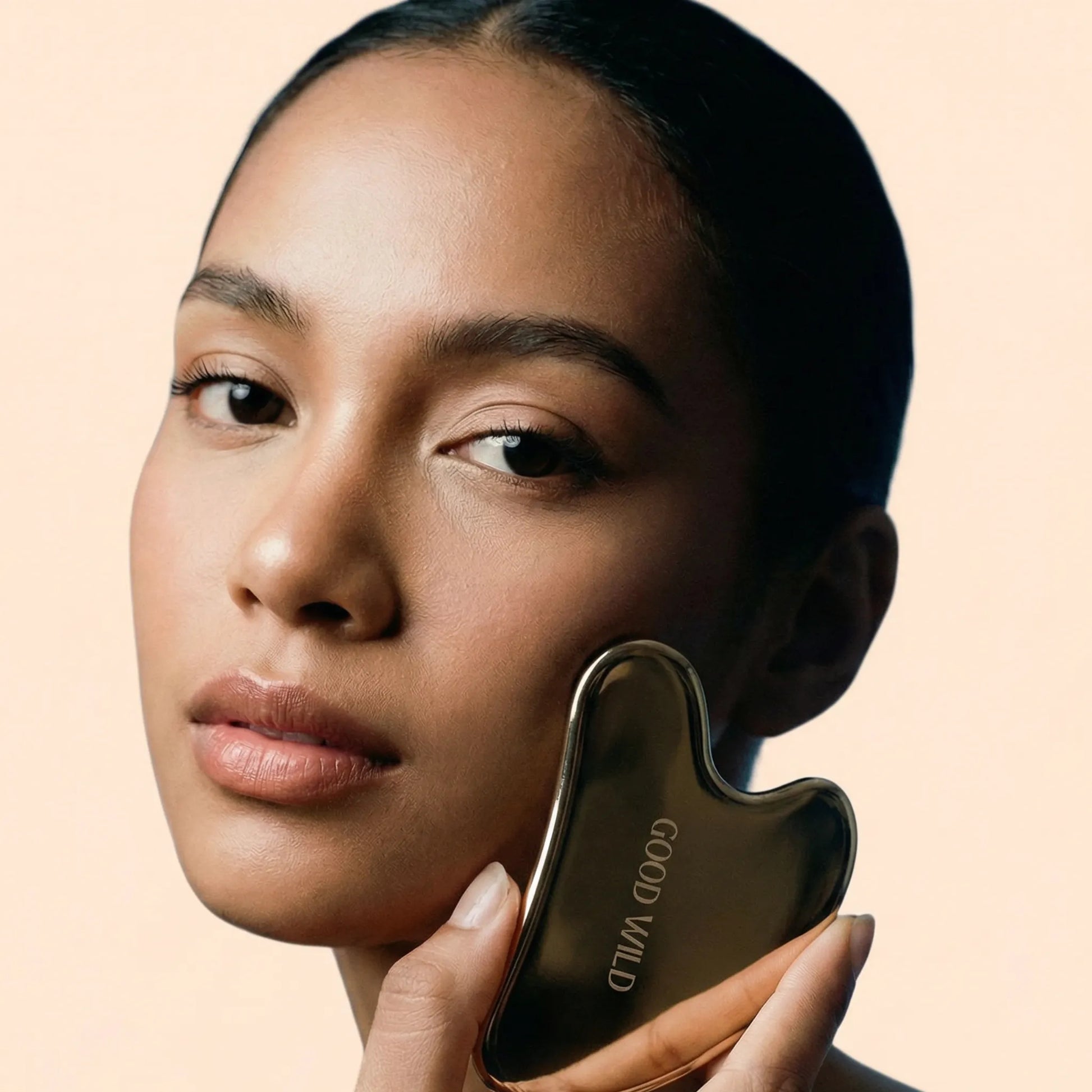 Woman holding a gold facial tool against her cheek on a beige background